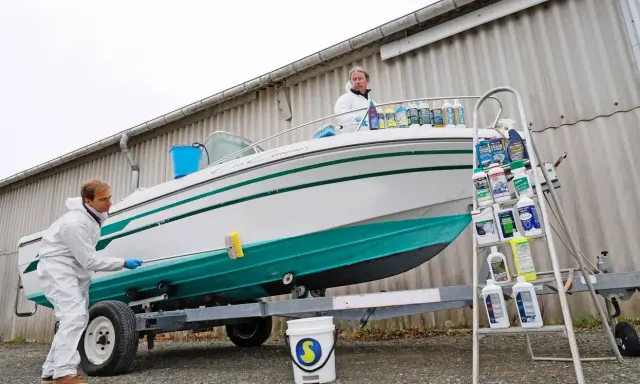 Un homme en combinaison blanche nettoie le flanc d'un bateau blanc et vert sur une remorque. Le nettoyage du pont du bateau est en cours.