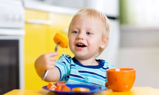 Niño rubio con tenedor y zanahoria, aprendiendo a comer solo. ¡Ayudame a hacerlo por mi mismo!