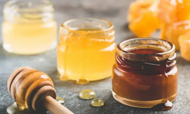 Trois pots de miel aux teintes variées, du clair au foncé, et une cuillère en bois dégoulinante. Quel miel pour pain d'épices ? Le choix est là !