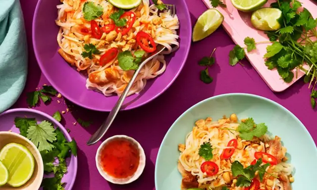 Två skålar med kyckling pad thai, toppad med chili, koriander och jordnötter. Limeklyftor och en liten skål med chilisås finns bredvid.