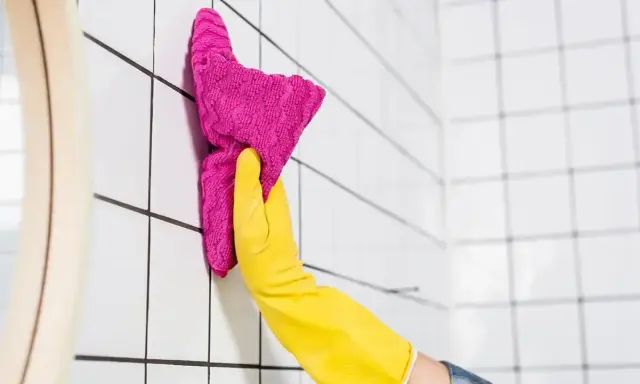 Ręka w żółtej rękawiczce czyści białe płytki różową ściereczką. To kluczowy krok, by dowiedzieć się, czym odtłuścić płytki przed malowaniem.