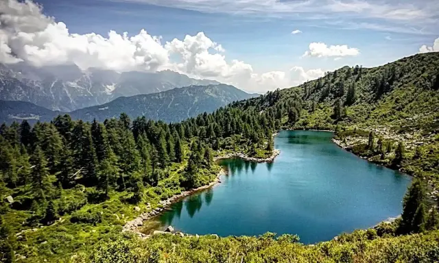 Laghi di San Giuliano: un'oasi alpina con acque turchesi, circondata da fitti boschi di conifere e montagne maestose sotto un cielo azzurro con nuvole bianche.