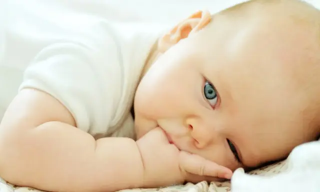 Niemowlę z niebieskimi oczami leży na boku, wkładając rączkę do ust. To uroczy moment, od kiedy niemowlę wkłada rączki do ust, odkrywając świat.