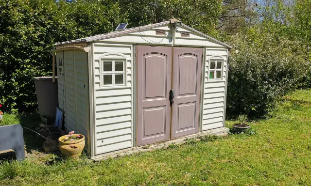 Un abri de jardin en plastique beige, avec des fenêtres et une double porte, est prêt pour son aménagement. Un petit panneau solaire est installé sur le toit.