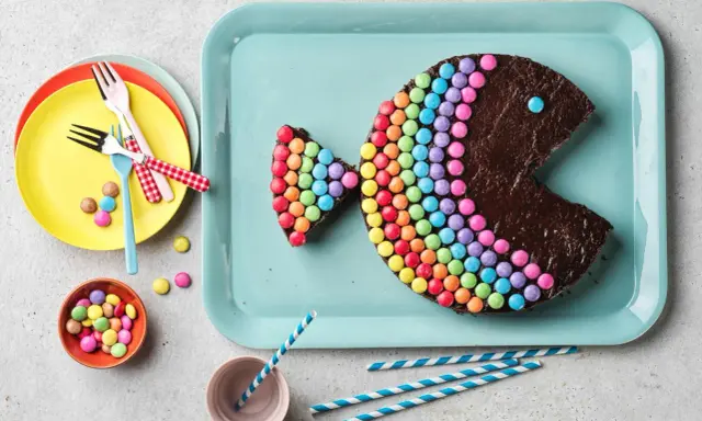 Gâteau en forme de poisson, décoré de Smarties formant un arc-en-ciel. Un œil bleu et une bouche ouverte complètent ce délice coloré.