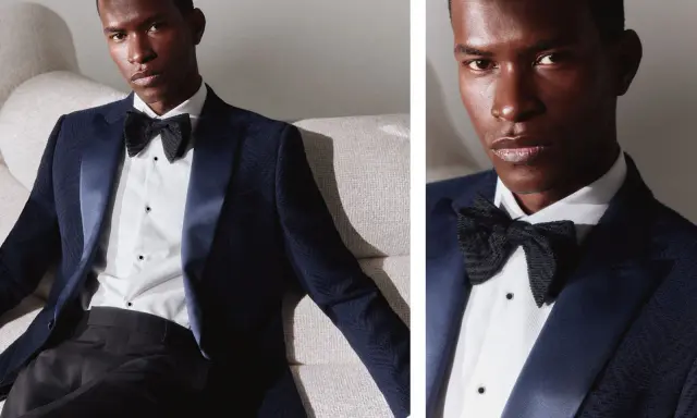 A man in a dark blue dinner jacket vs tuxedo, with a satin lapel and black bow tie, poses on a textured sofa.