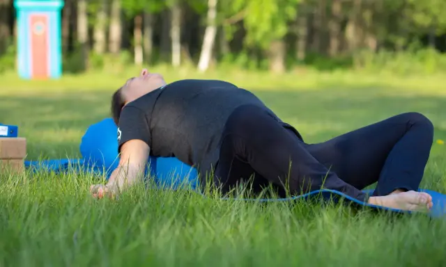 Person ligger på en blå yogarulle i gräset, med böjda knän och armarna utåt. En lugn stund med yin yoga utomhus.