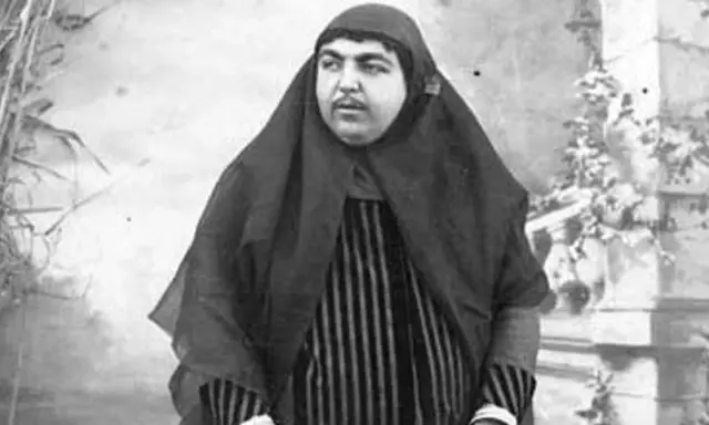 Zahra Khanom Tadj es-Saltaneh, księżniczka perska, w czarnym hidżabie i pasiastym stroju, pozuje na tle ogrodu.