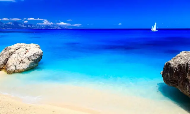 Spiaggia con rocce e mare cristallino, un veliero solca le acque turchesi. Potrebbe essere il mare più bello in Italia.