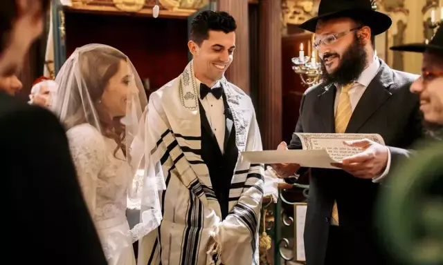 Novios sonríen durante su **matrimonio judío**. El novio lleva un talit y el rabino lee un documento.