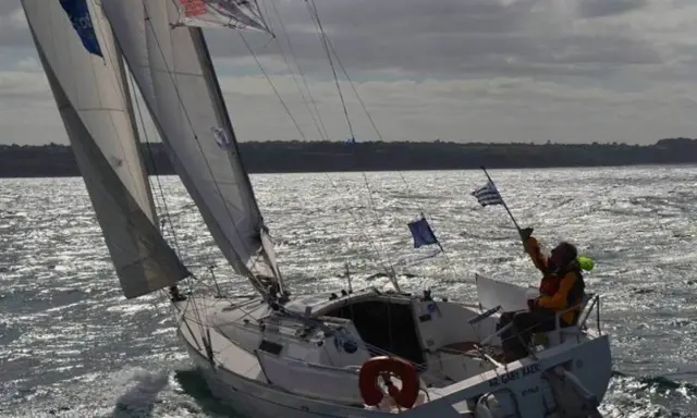Voilier naviguant par gros temps, le skipper tient un drapeau. La fiche technique du premier 260 est visible sur la coque.