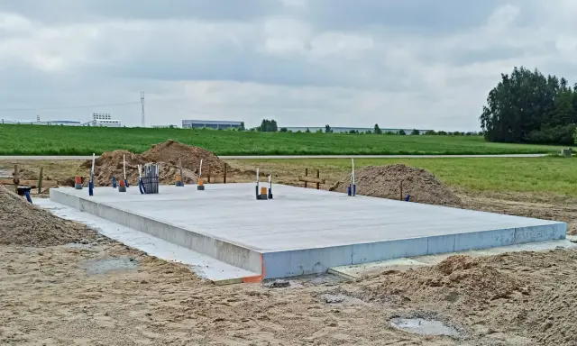 Gotowe fundamenty krok po kroku. Płyta betonowa na budowie, otoczona piaskiem i zielenią. W tle widać budynki przemysłowe.