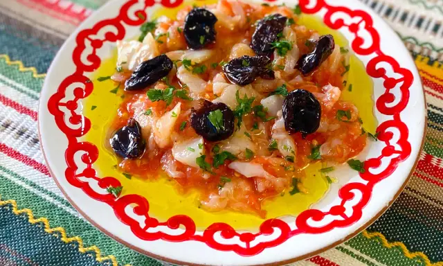 Esqueixada de bacalao con aceitunas negras y perejil, servida en un plato blanco con borde rojo sobre un mantel colorido.
