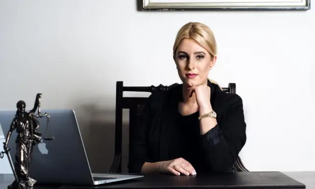 Kobieta w czarnej marynarce siedzi przy biurku z laptopem i figurką Temidy.