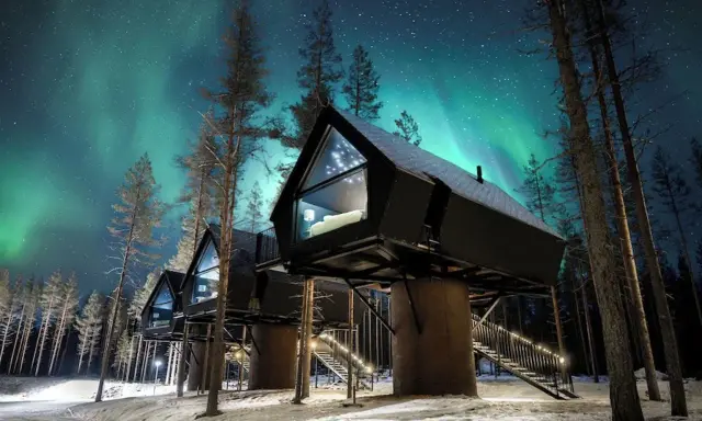 Hus på styltor i snöig skog under norrsken. En **arctic road** leder fram till stugorna, upplysta av stjärnor och lampor.