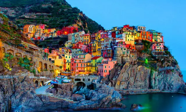 Case colorate aggrappate alla scogliera, un incantevole scorcio di quali sono le 5 Terre, con barche ormeggiate e il mare blu.