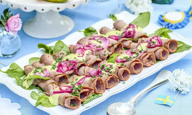 Rullad rostbiff med grön sås och rödlök, perfekt som enkel buffémat till många. Serveras på en vit bricka med gröna blad.