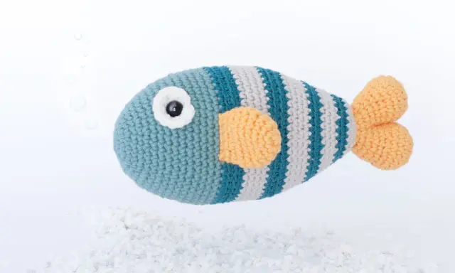 Un pez amigurumi a rayas azules y grises con aletas naranjas, flotando sobre guijarros blancos. ¡Encuentra aquí peces amigurumi patrón gratis!