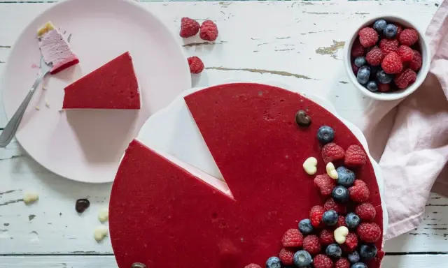 Ein köstlicher no bake Himbeer Cheesecake, dekoriert mit frischen Beeren und Schokoharz. Ein Stück ist bereits auf einem Teller.
