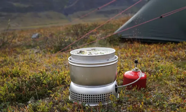 Matlagning i fjällen med ett multifuelkök. En kastrull med mat värms upp bredvid ett tält.