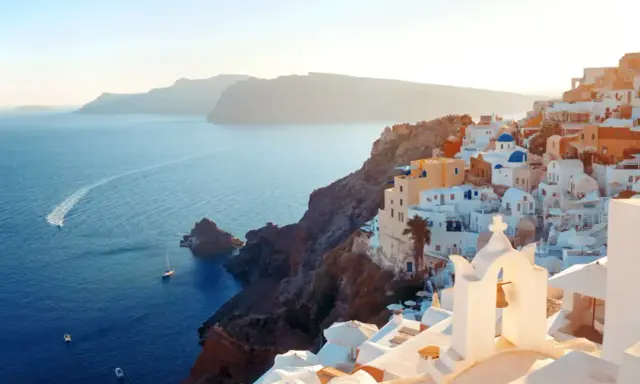 Traumhafter Blick auf die weißen Häuser und blauen Kuppeln von Santorini. Das Meer glitzert, Boote fahren vorbei. Ein perfekter Santorini Strand Urlaub.