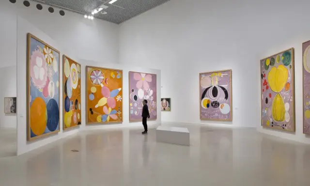En person betraktar färgglada, abstrakta målningar av Hilma af Klint i ett ljust galleri.