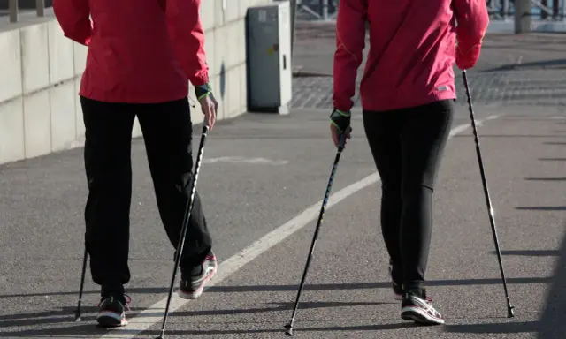 Dwie osoby w czerwonych kurtkach i czarnych spodniach maszerują z kijki do nordic walking.