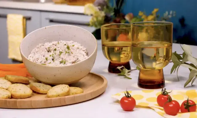 Un bol de rillettes de thon crémeuses, garni de ciboulette, servi avec des crackers, des bâtonnets de carottes et de céleri. Deux verres de vin blanc complètent cette scène gourmande.
