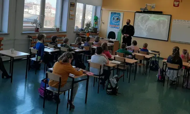 Dzieci słuchają lekcji o bezpieczeństwie w szkole. Nauczycielka i policjant prowadzą zajęcia.