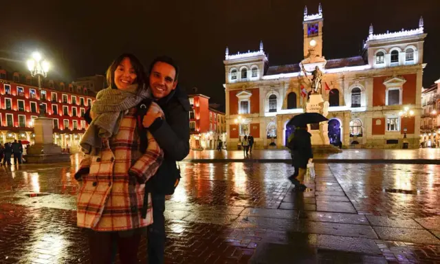 Pareja sonriente en la Plaza Mayor de Valladolid, disfrutando de las atracciones nocturnas y el reflejo de las luces en el suelo mojado.