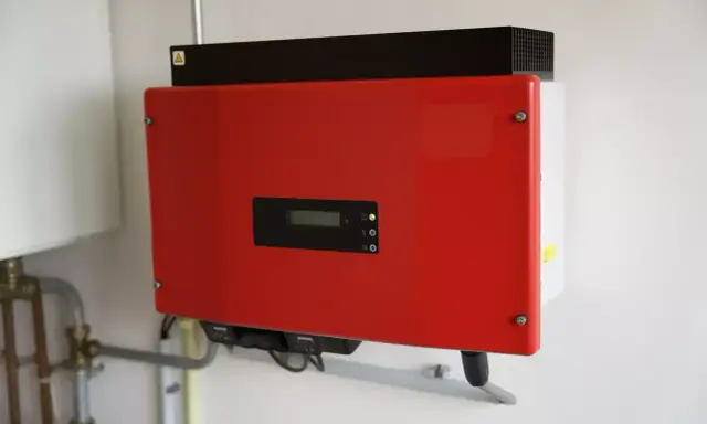 Röd kostal växelriktare monterad på en vit vägg. Enheten har en liten display och indikatorlampor.