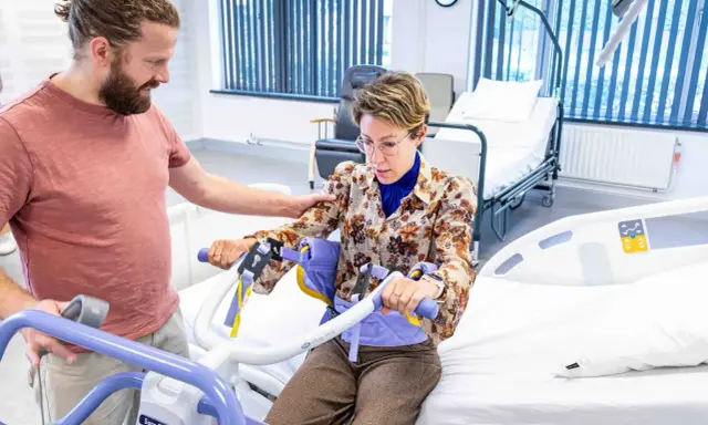 Un homme aide une femme à s'asseoir sur un lève-personne, illustrant l'ergonomie infirmier pour un transfert sécurisé.