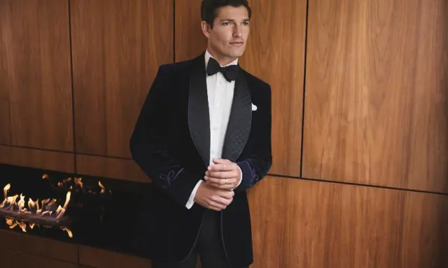 A man in a sophisticated navy velvet tuxedo, embodying perfect black tie attire, stands by a modern fireplace.
