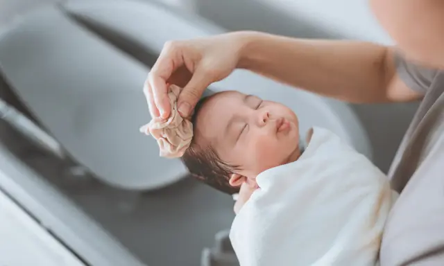 Bain d'un nouveau-né : une main nettoie délicatement la tête du bébé avec une petite serviette. Une scène paisible qui rappelle la liste produits toilette bébé naissance.