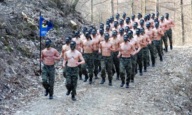 Żołnierze w kamuflażu biegają w lesie, niektórzy z zielonym malowaniem twarzy i flagą.