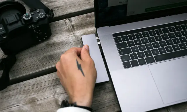 Dłoń z tatuażem pioruna podłącza adapter do laptopa, obok leży aparat. Idealny zestaw, jaki laptop do obróbki zdjęć?