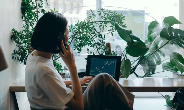 Kobieta w białej koszulce rozmawia przez telefon, pracując na laptopie z wykresem giełdowym. To jej wolny zawód, który pozwala jej pracować w otoczeniu zieleni.