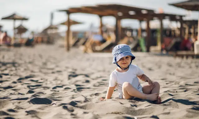 Uroczy maluch w pasiastym kapelusiku siedzi na piaszczystej plaży. Idealne miejsce, gdzie na wakacje z rocznym dzieckiem.