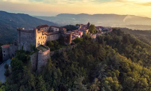 Antico castello medievale su una collina boscosa, uno dei tanti castelli da visitare in Toscana, con un villaggio arroccato e vista sulla valle al tramonto.
