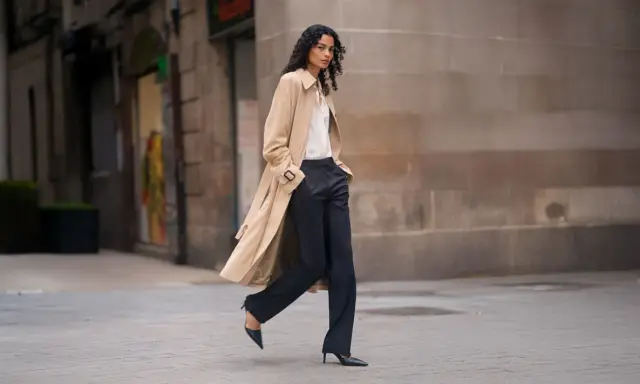 A woman walks confidently in dark palazzo pants and a trench coat, proving they are still in style.