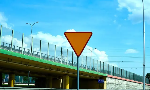 Pomarańczowy znak "ustąp pierwszeństwa" ostrzega przed wjazdem na drogę podporządkowaną. Pod wiaduktem widać drogę i słupy oświetleniowe.