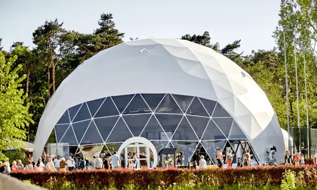 Una gran cúpula estructura blanca, con ventanas triangulares, se alza en un día soleado. Gente pasea frente a ella, rodeada de árboles verdes.