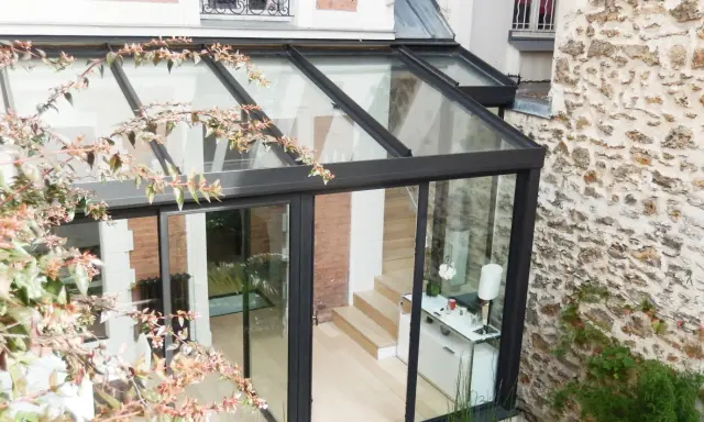 Une pergola transformée en véranda moderne, avec une structure en verre et métal, s'intégrant à une façade en pierre et brique.