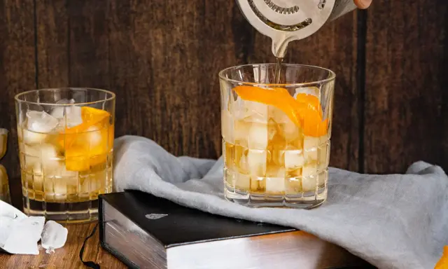 Ein Old Fashioned wird zubereitet, Eiswürfel und Orangenschale zieren das Glas. Ein altes Rezept wird lebendig.