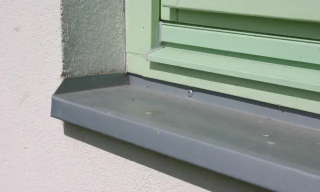 Szary parapet zewnętrzny zamontowany przy oknie z zielonymi okiennicami. Idealny moment, kiedy montować parapety zewnętrzne, to po ociepleniu elewacji.