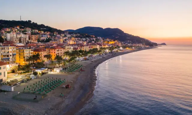 Alba sulla spiaggia finale ligure, con file di ombrelloni verdi e palme che si affacciano sul mare calmo.