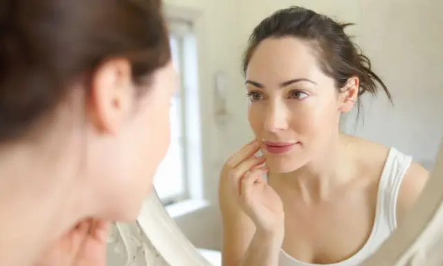 Une femme examine son visage dans un miroir, touchant sa joue. Sa peau semble très fine, peut-être signe d'une carence.