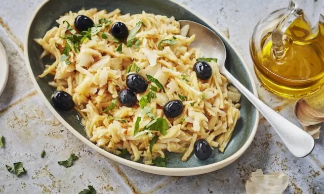 Un plat de bacalhau à brás, effiloché et garni d'olives noires et de persil frais. Une touche d'huile d'olive et d'ail est visible à côté.
