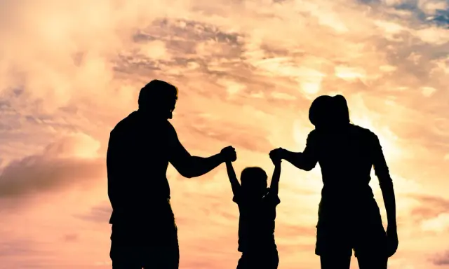 Siluetas de una familia jugando bajo un cielo anaranjado. Un niño es levantado por sus padres, un ejemplo de aprendizaje social y emocional.