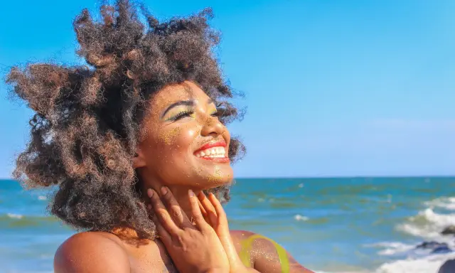 Donna con capelli afro e trucco scintillante si gode il sole in spiaggia. Sembra che ci si abbronza con il trucco, creando un look festoso.